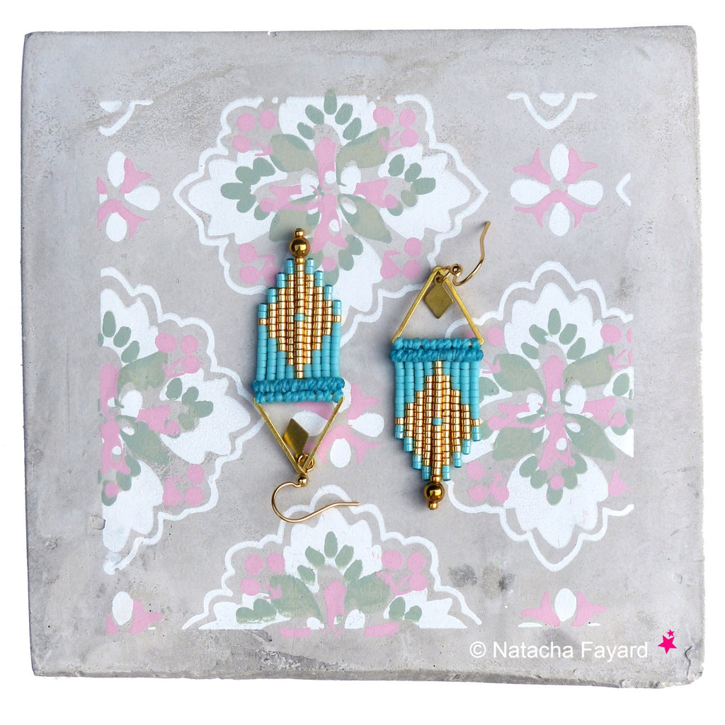 Peut inclure: Une paire de boucles d'oreilles en perles dorées et turquoise avec un motif géométrique. Les boucles d'oreilles sont suspendues à un fond gris à motif floral.