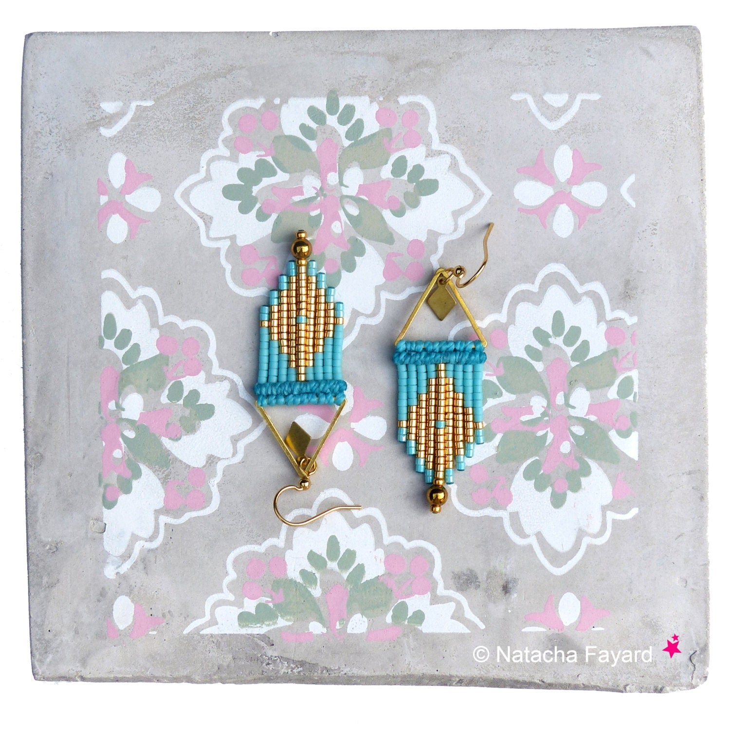 Peut inclure: Une paire de boucles d'oreilles en perles dorées et turquoise avec un motif géométrique. Les boucles d'oreilles sont suspendues à un fond gris à motif floral.