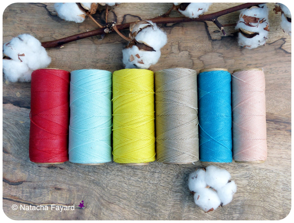 Peut inclure: Six bobines de fil de coton de différentes couleurs, dont rouge, bleu, jaune, beige, turquoise et rose. Les bobines sont disposées en rangée sur une surface en bois avec des boules de coton de chaque côté.