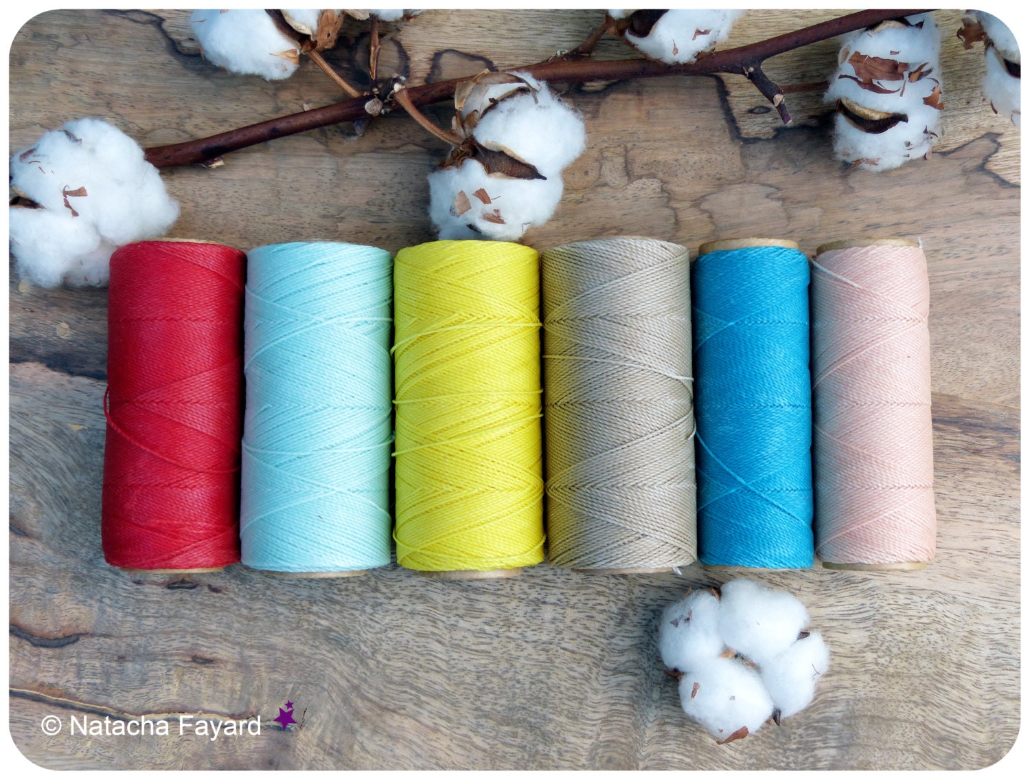Peut inclure: Six bobines de fil de coton de différentes couleurs, dont rouge, bleu, jaune, beige, turquoise et rose. Les bobines sont disposées en rangée sur une surface en bois avec des boules de coton de chaque côté.