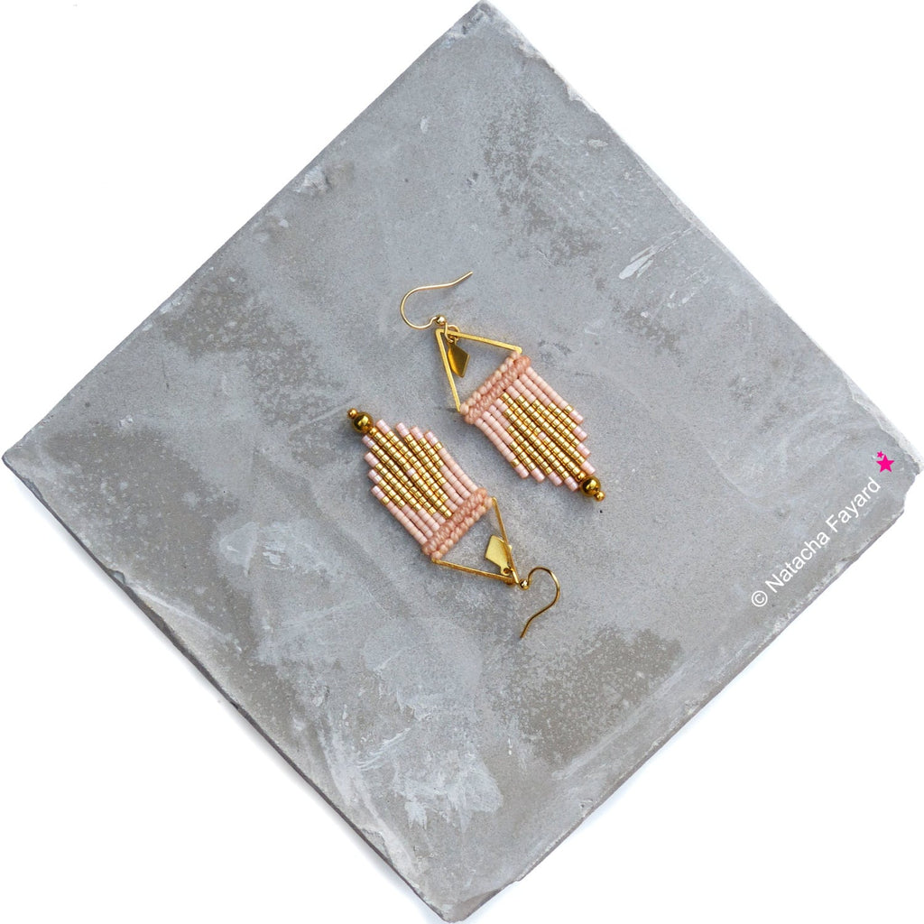 Peut inclure: Une paire de boucles d'oreilles en perles dorées et roses avec un design géométrique. Les boucles d'oreilles sont suspendues à une surface grise.