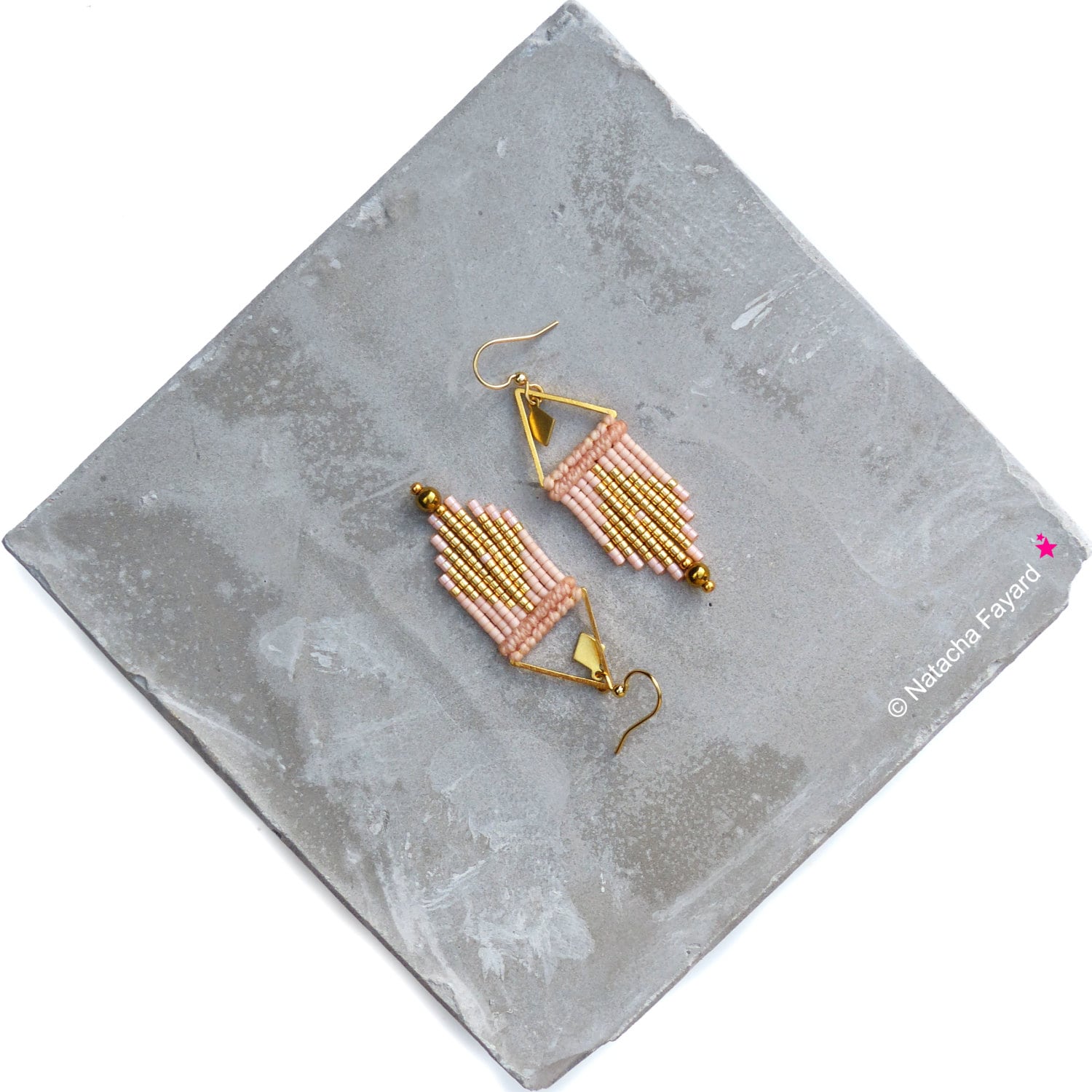 Peut inclure: Une paire de boucles d'oreilles en perles dorées et roses avec un design géométrique. Les boucles d'oreilles sont suspendues à une surface grise.