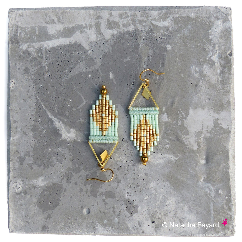Peut inclure: Une paire de boucles d'oreilles géométriques en perles dorées et vert clair de forme triangulaire. Les boucles d'oreilles sont suspendues à une surface en béton.
