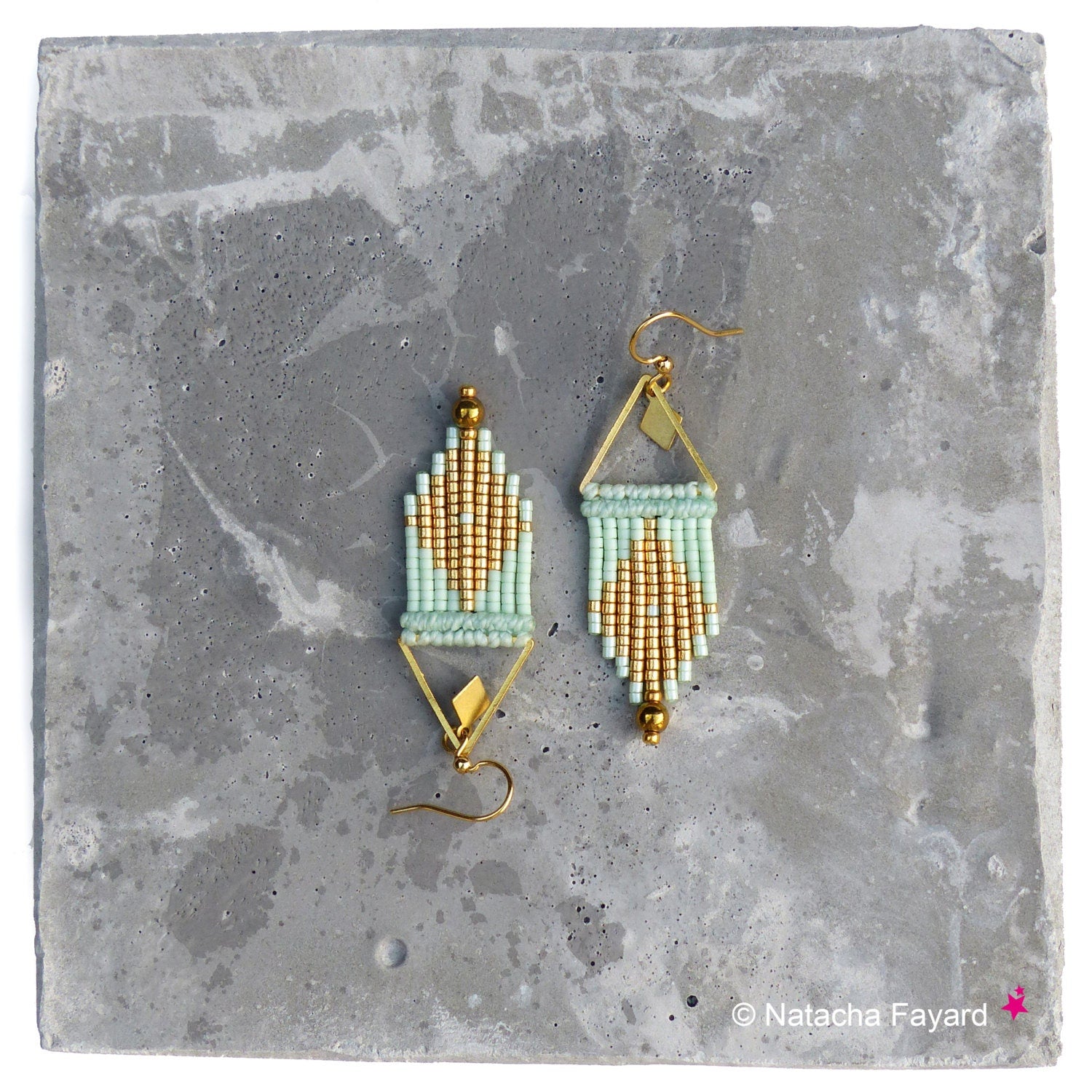 Peut inclure: Une paire de boucles d'oreilles géométriques en perles dorées et vert clair de forme triangulaire. Les boucles d'oreilles sont suspendues à une surface en béton.