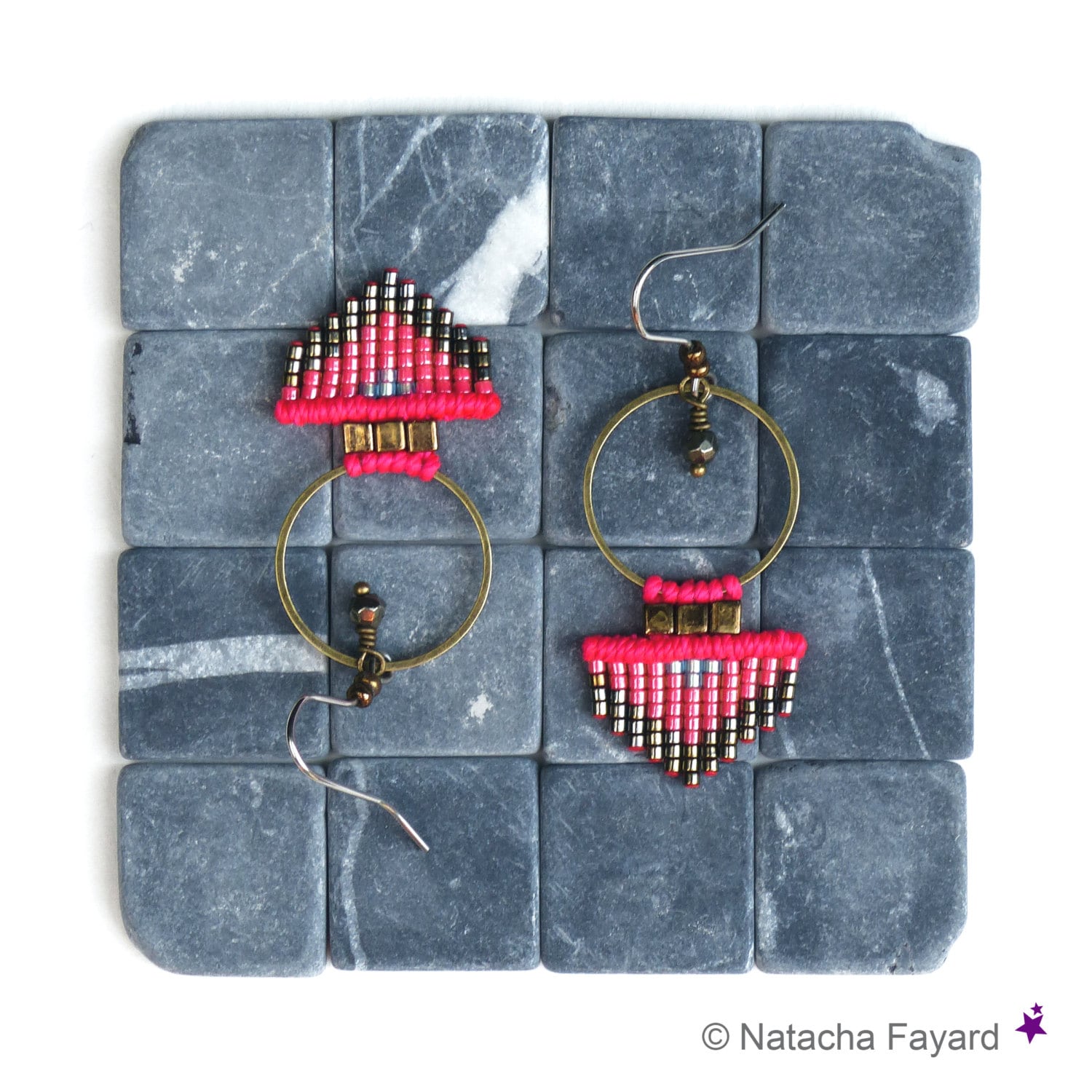 Peut inclure: Une paire de boucles d'oreilles en perles roses et argentées avec un motif géométrique. Les boucles d'oreilles sont suspendues à une surface carrelée grise.