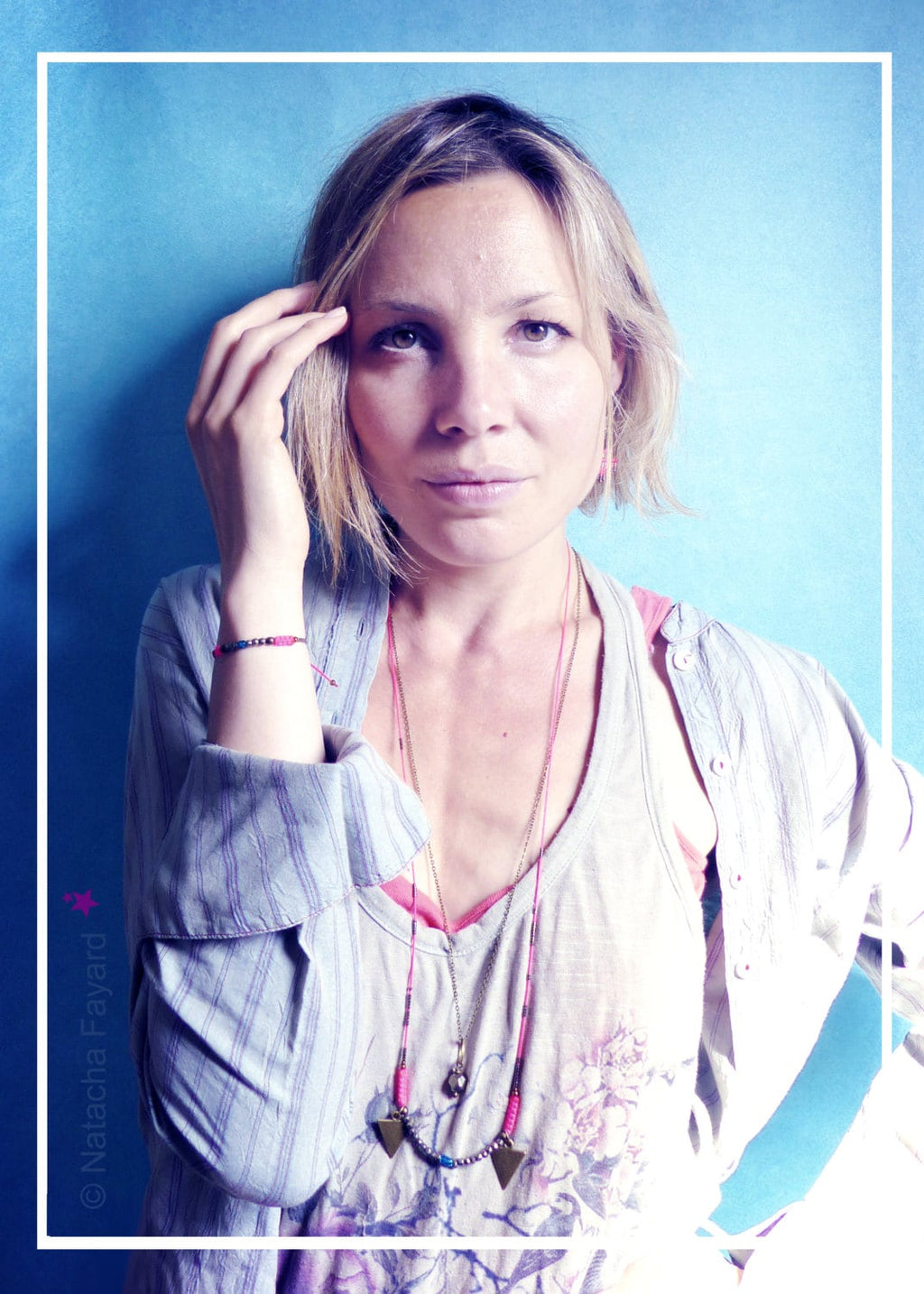 Peut inclure: Une femme aux cheveux blonds courts porte une chemise gris clair avec un collier superposé rose et bleu. Le collier a deux pendentifs triangulaires. Elle porte un bracelet rose et bleu au poignet gauche.