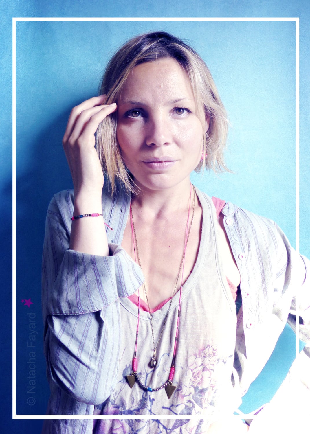 Peut inclure: Une femme aux cheveux blonds courts porte une chemise gris clair avec un collier superposé rose et bleu. Le collier a deux pendentifs triangulaires. Elle porte un bracelet rose et bleu au poignet gauche.