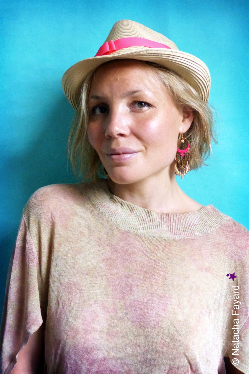 Peut inclure: Une femme aux cheveux blonds porte un chapeau de paille brun clair avec un ruban rose et une paire de boucles d'oreilles en perles dorées et roses. Elle porte une chemise tie-dye brun clair, rose et blanc.