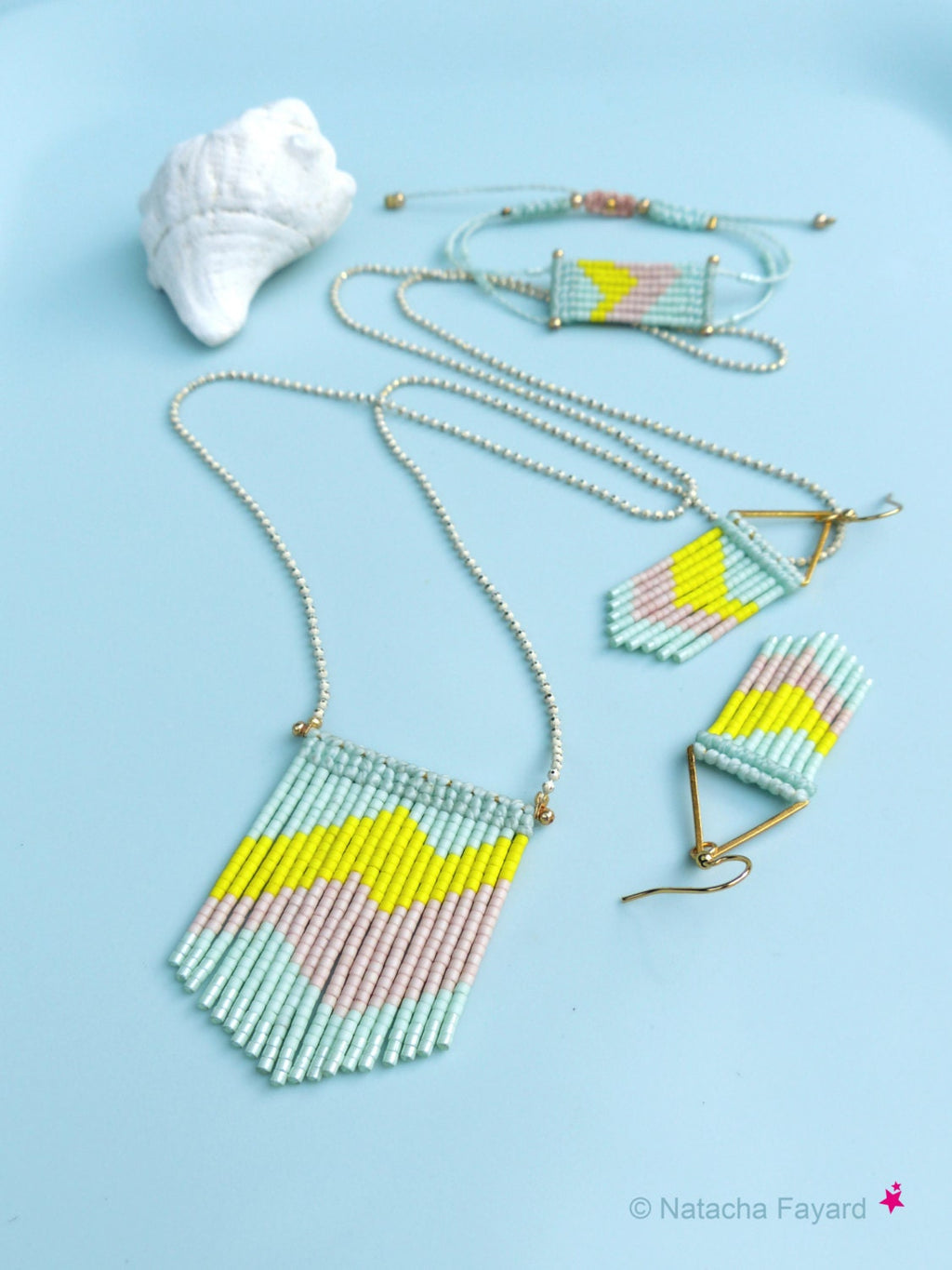 Peut inclure: Un ensemble de trois bijoux en perles avec un motif géométrique. Le collier, le bracelet et les boucles d'oreilles sont faits de perles bleu clair, rose et jaune. Les bijoux sont sur un fond bleu clair.
