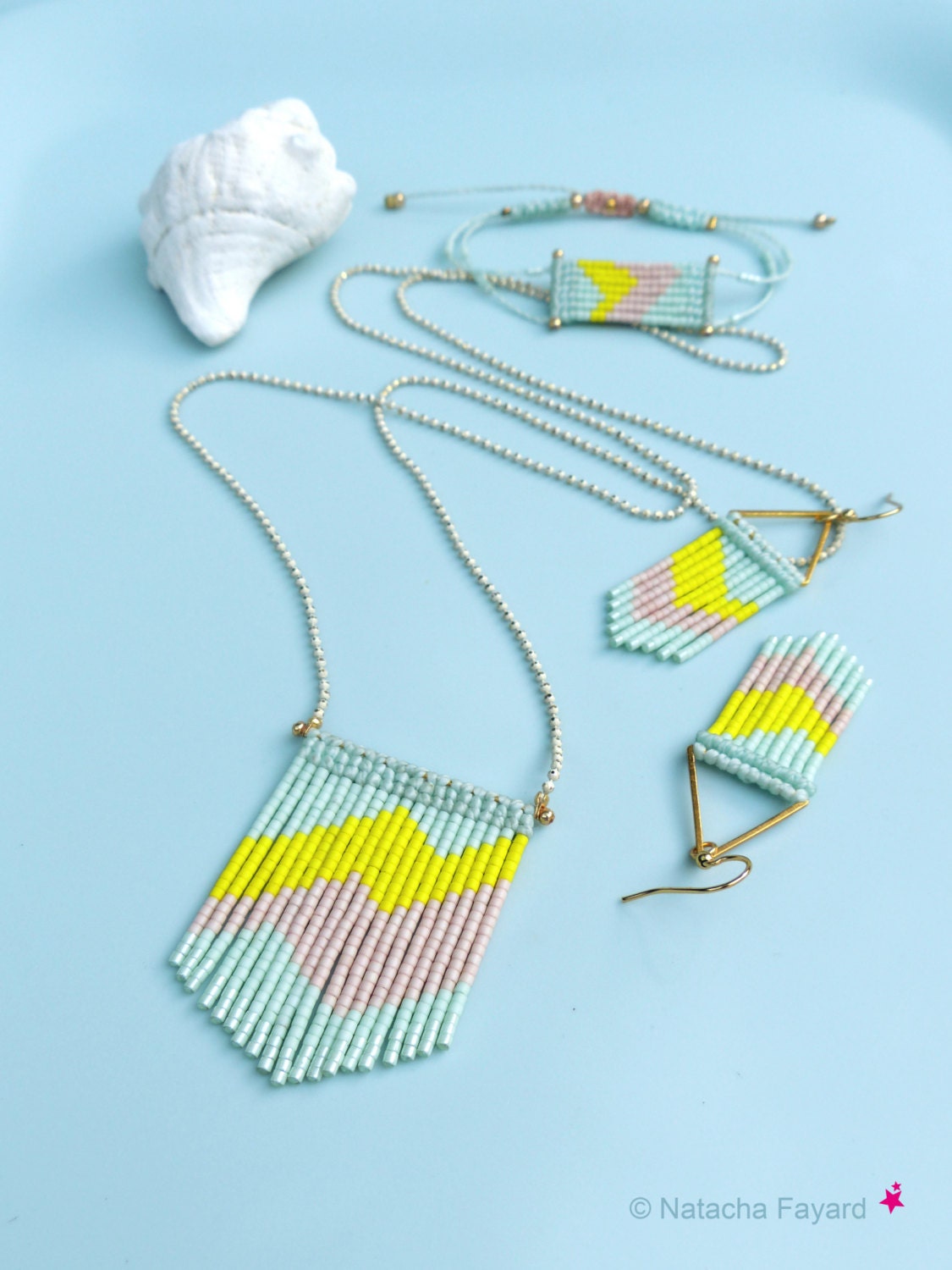 Peut inclure: Un ensemble de trois bijoux en perles avec un motif géométrique. Le collier, le bracelet et les boucles d'oreilles sont faits de perles bleu clair, rose et jaune. Les bijoux sont sur un fond bleu clair.