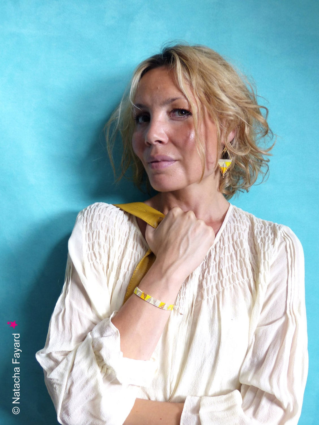 Peut inclure: Une femme portant une blouse blanche avec un bracelet en perles jaunes et des boucles d'oreilles en perles jaunes. Elle est debout devant un fond bleu.