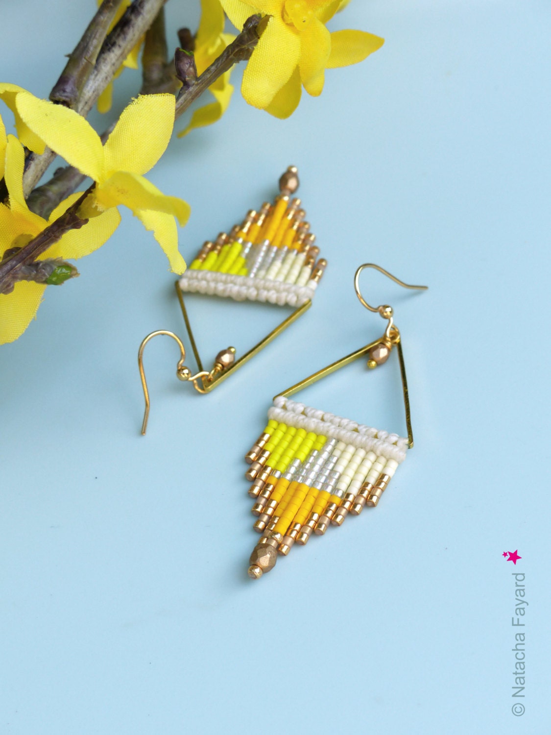 Peut inclure: Une paire de boucles d'oreilles triangulaires dorées avec des perles jaunes, blanches et argentées. Les boucles d'oreilles sont suspendues sur un fond bleu clair.