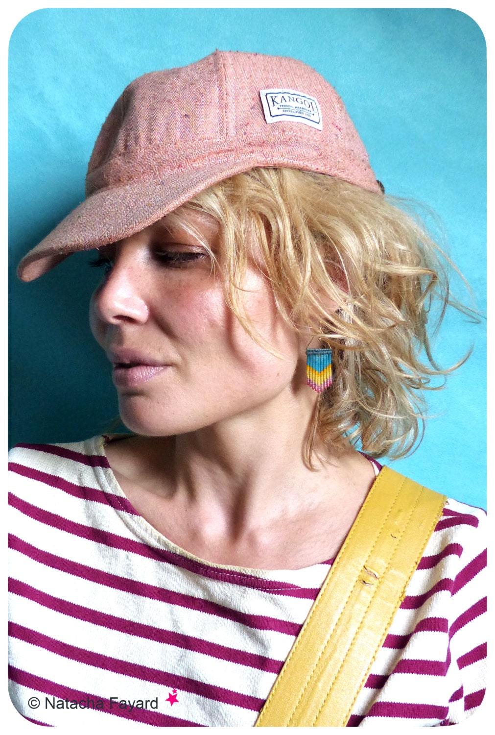 Peut inclure: Une femme portant une casquette de baseball rose et blanche mouchetée avec le mot "Kangol" dessus. Elle porte une chemise rayée avec une bretelle jaune et a une boucle d'oreille en perles à l'oreille droite.