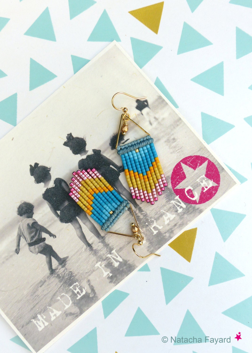 Peut inclure: Une paire de boucles d'oreilles en triangle doré avec des franges de perles bleues, jaunes et roses. Les boucles d'oreilles sont présentées sur une carte postale vintage avec le texte "Made in France".