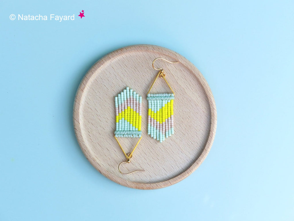 Peut inclure: Une paire de boucles d'oreilles en perles géométriques avec un motif bleu clair, jaune et rose. Les boucles d'oreilles sont suspendues à des anneaux dorés en forme de triangle.