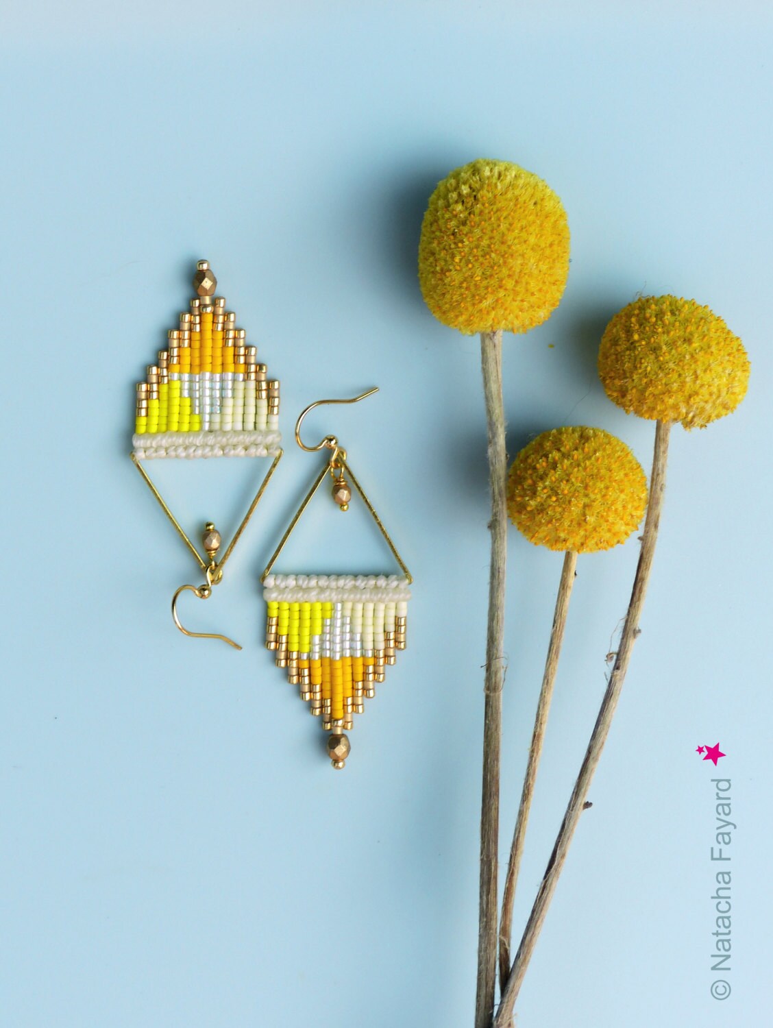 Peut inclure: Une paire de boucles d'oreilles triangulaires dorées avec des franges de perles jaunes, blanches et dorées. Les boucles d'oreilles sont photographiées sur un fond bleu clair avec des fleurs jaunes séchées.