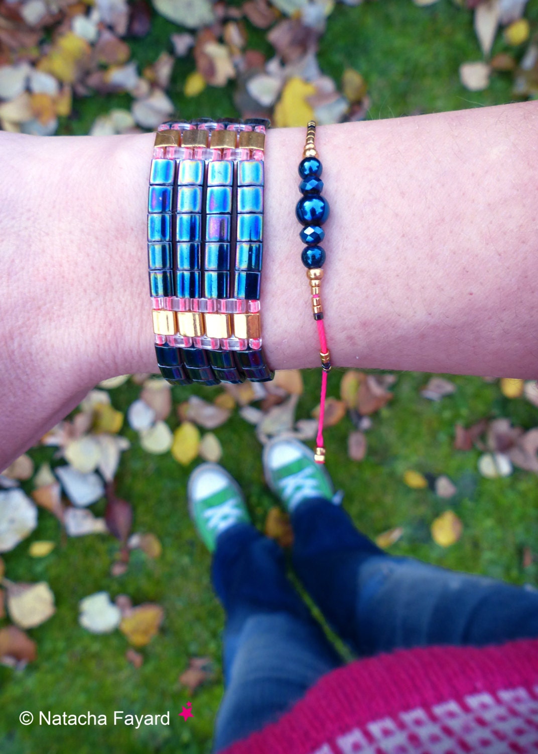 Peut inclure: Gros plan sur le poignet d'une personne portant deux bracelets. Le premier bracelet est un bracelet multi-couches, bleu irisé et or. Le deuxième bracelet est une simple chaîne dorée avec des perles bleu foncé. Les bracelets sont portés sur un bras nu sur fond d'herbe verte et de feuilles mortes.