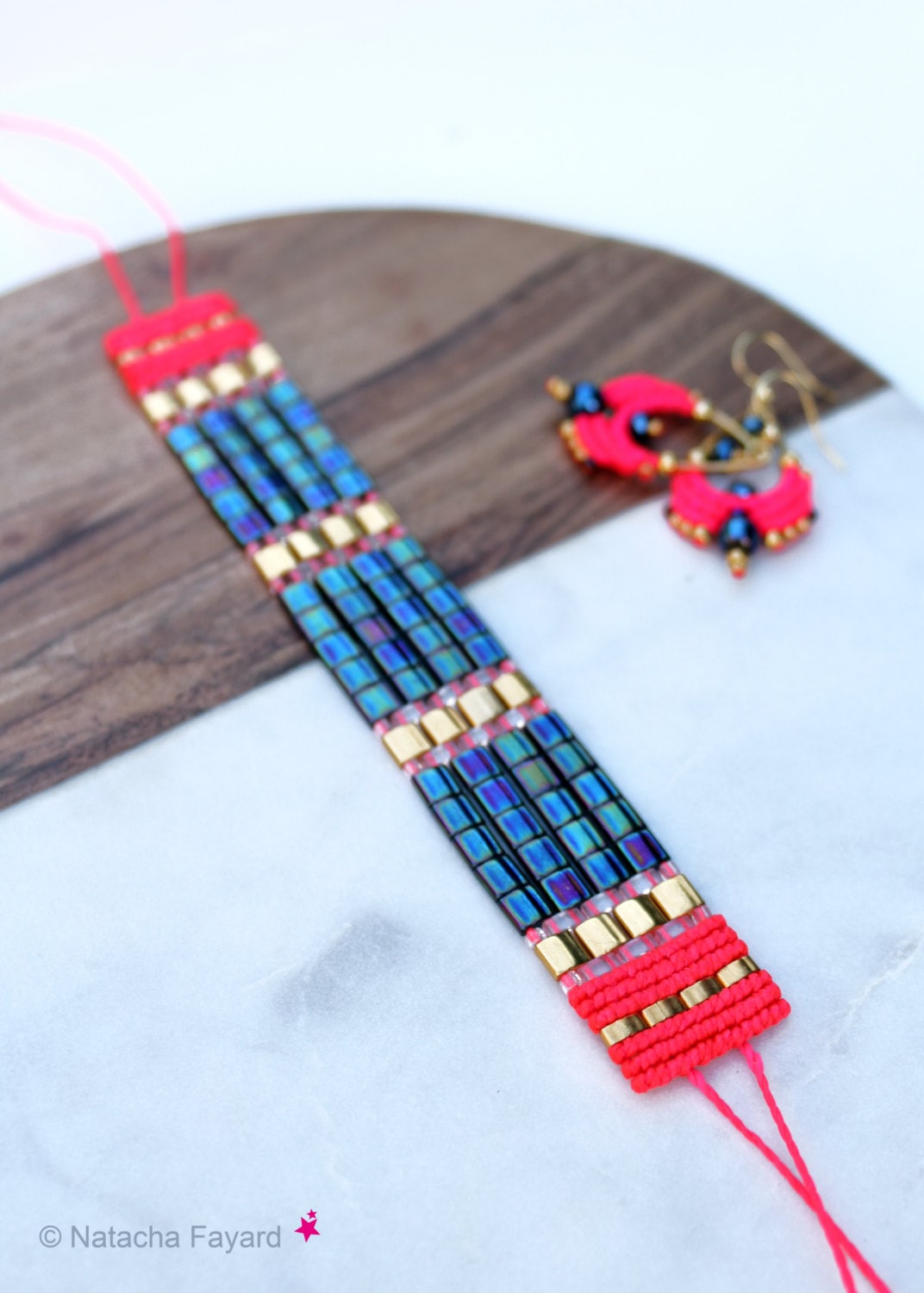 Peut inclure: Un bracelet en perles rose et or avec des perles carrées bleu et noir irisées. Le bracelet est sur une surface en bois avec une paire de boucles d'oreilles en perles rose et noir.