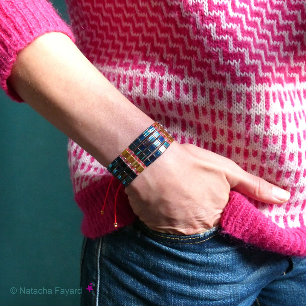 Peut inclure: Un bracelet en perles bleues, dorées et roses avec une fermeture à cordon rouge. Le bracelet est porté au poignet.