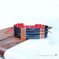 Peut inclure: Un bracelet multi-rangs avec des perles carrées bleu et vert irisées, des perles dorées et du fil rose.