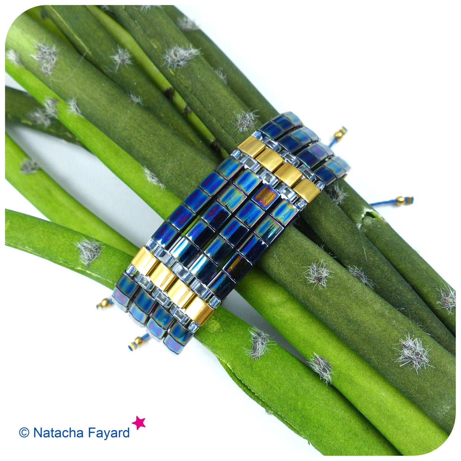 Peut inclure: Un bracelet multi-rangs composé de perles carrées bleu irisé et dorées. Le bracelet est enroulé autour de tiges vertes.