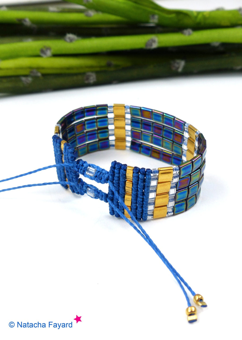 Peut inclure: Un bracelet bleu et or avec une fermeture à nœud coulissant. Le bracelet est composé de perles carrées bleues irisées et dorées et d'un cordon bleu.