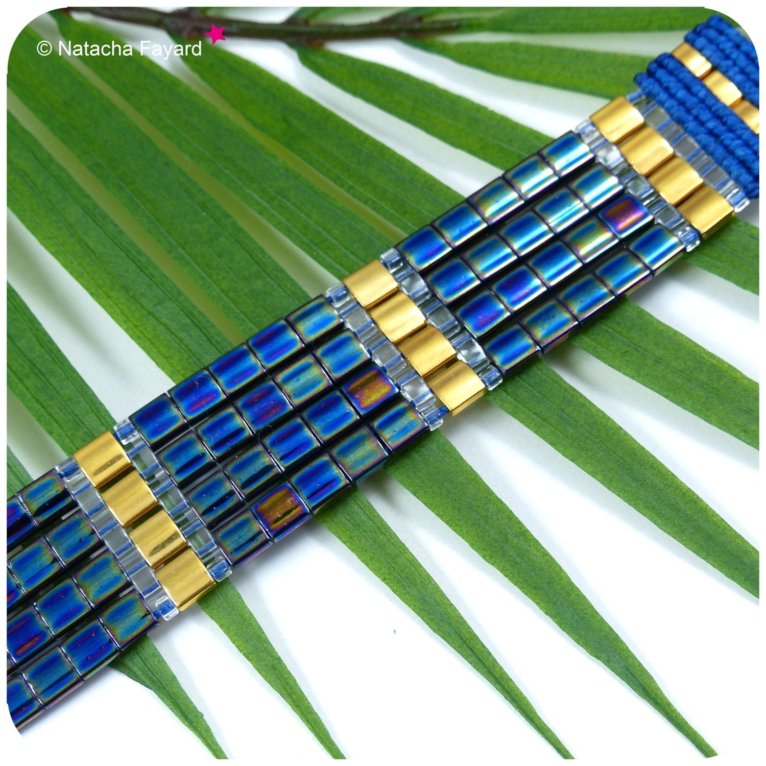 Peut inclure: Un bracelet multi-colore en gros plan avec des perles bleues, dorées et argentées. Le bracelet est composé de petites perles carrées et est posé sur une feuille verte.