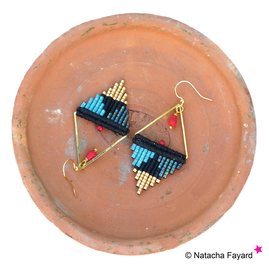 Peut inclure: Une paire de boucles d'oreilles triangulaires géométriques avec des perles noires, bleues et dorées. Les boucles d'oreilles ont des accents rouges et un fil doré.
