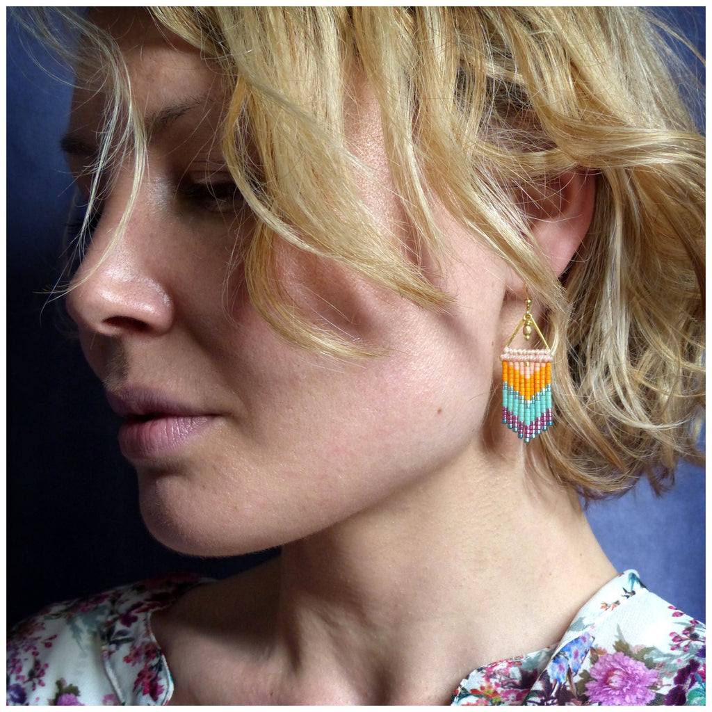 Peut inclure: Gros plan sur une boucle d'oreille perlée avec un dessus triangulaire doré et un motif vertical coloré. La boucle d'oreille présente des perles orange, turquoise et violettes. La boucle d'oreille est suspendue à un crochet doré.