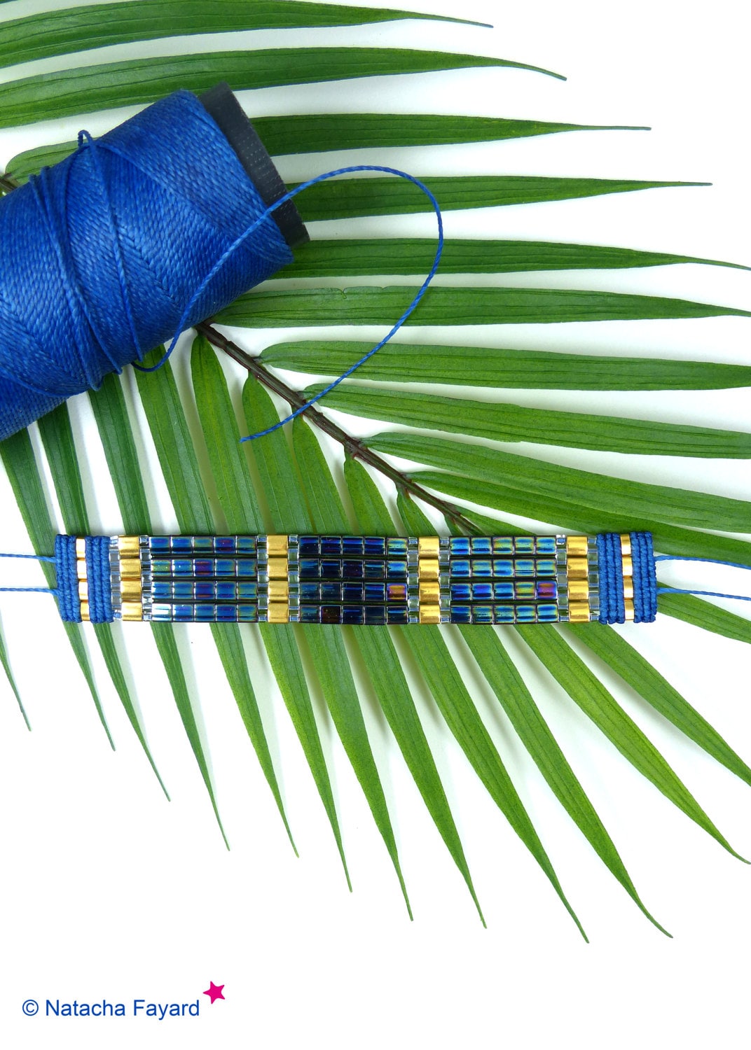 Peut inclure: Un bracelet en perles bleues et dorées avec un motif géométrique. Le bracelet est posé sur une feuille de palmier verte.
