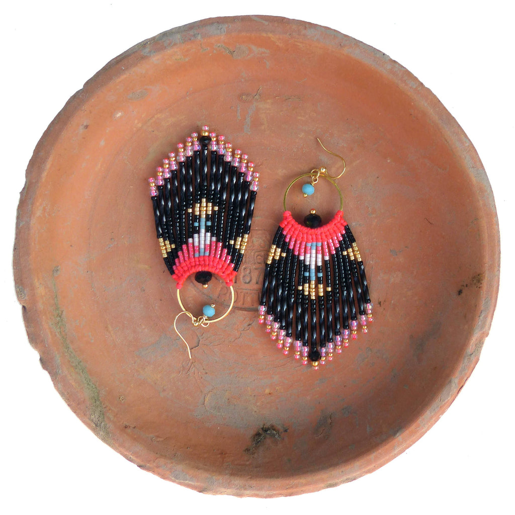 Peut inclure: Une paire de boucles d'oreilles en perles noires, roses et dorées avec un design en frange. Les boucles d'oreilles sont suspendues à une assiette en terre cuite.