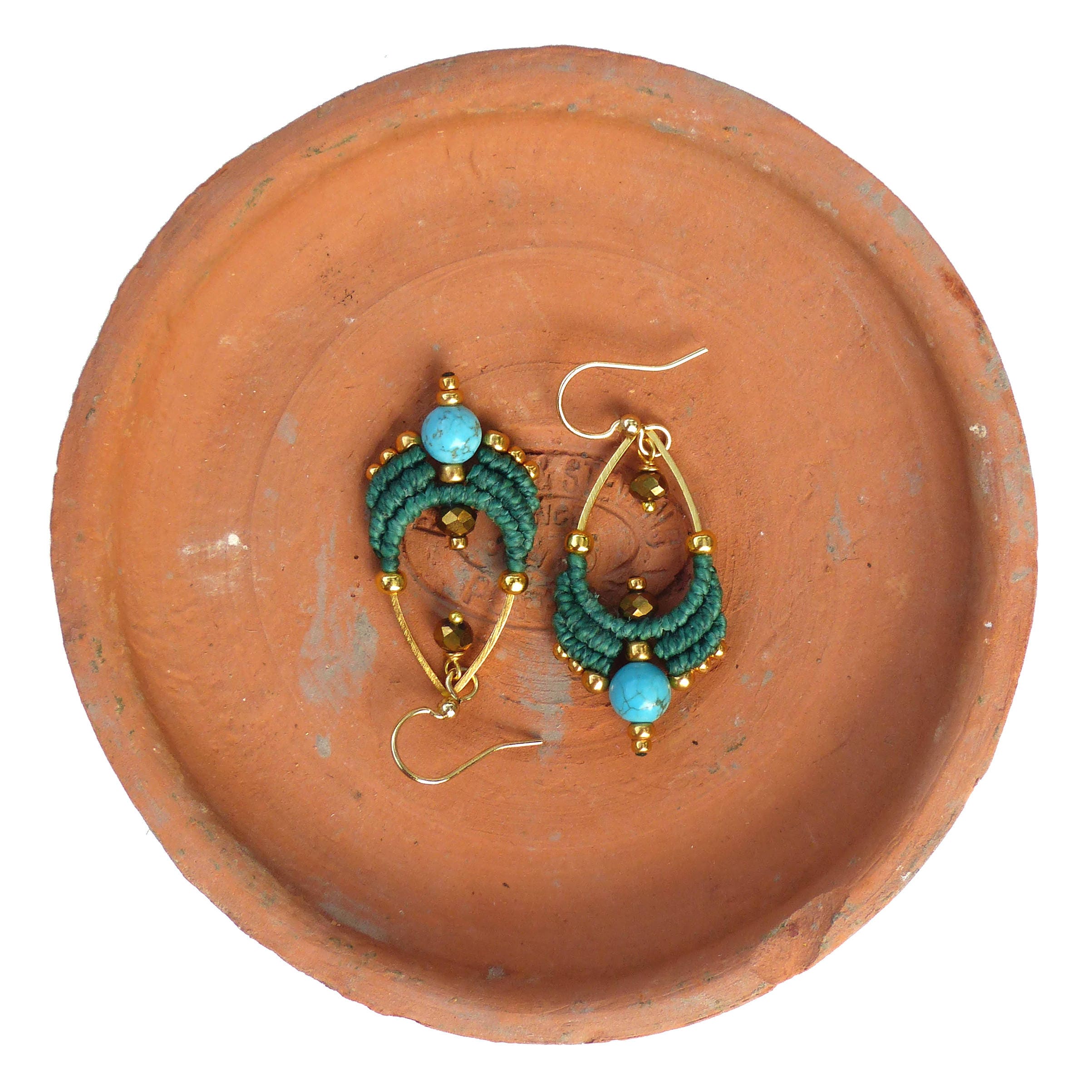 Peut inclure: Une paire de boucles d'oreilles vertes et dorées avec des pierres turquoise. Les boucles d'oreilles sont suspendues à une assiette en terre cuite.