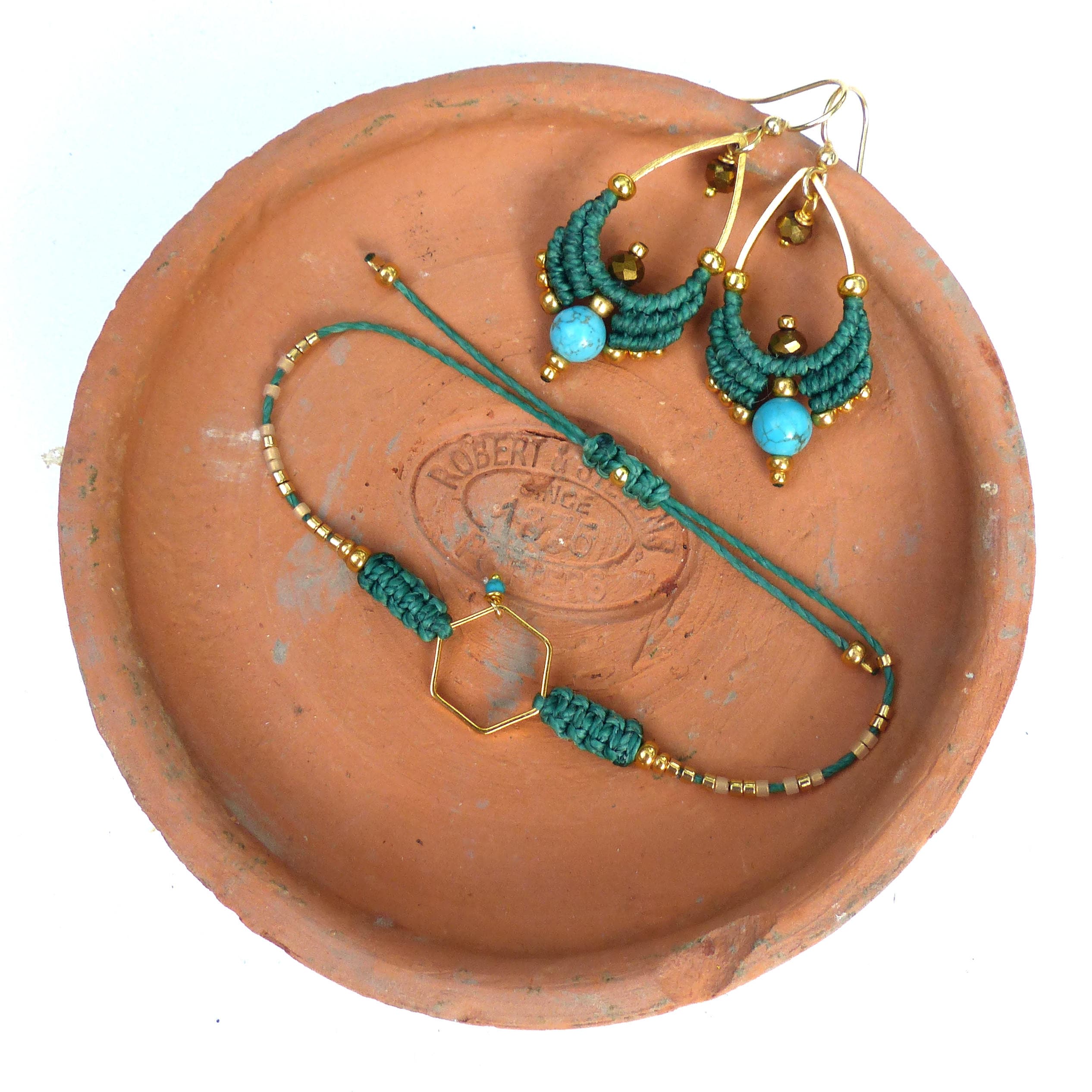Peut inclure: Un ensemble de boucles d'oreilles en macramé vert et or et un bracelet avec des perles turquoise. Les bijoux sont présentés sur une assiette en terre cuite.