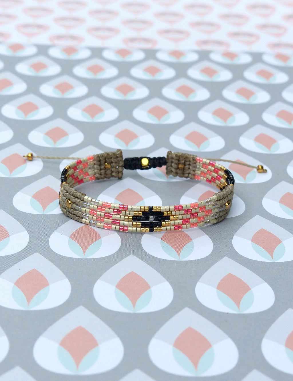 Peut inclure: Un bracelet tissé avec un motif géométrique. Le bracelet présente une base beige avec des rangées de perles roses, noires et dorées. Il est réglable avec des cordons beiges et des détails dorés.