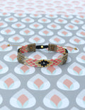 Peut inclure: Un bracelet tissé avec un motif géométrique. Le bracelet présente une base beige avec des rangées de perles roses, noires et dorées. Il est réglable avec des cordons beiges et des détails dorés.