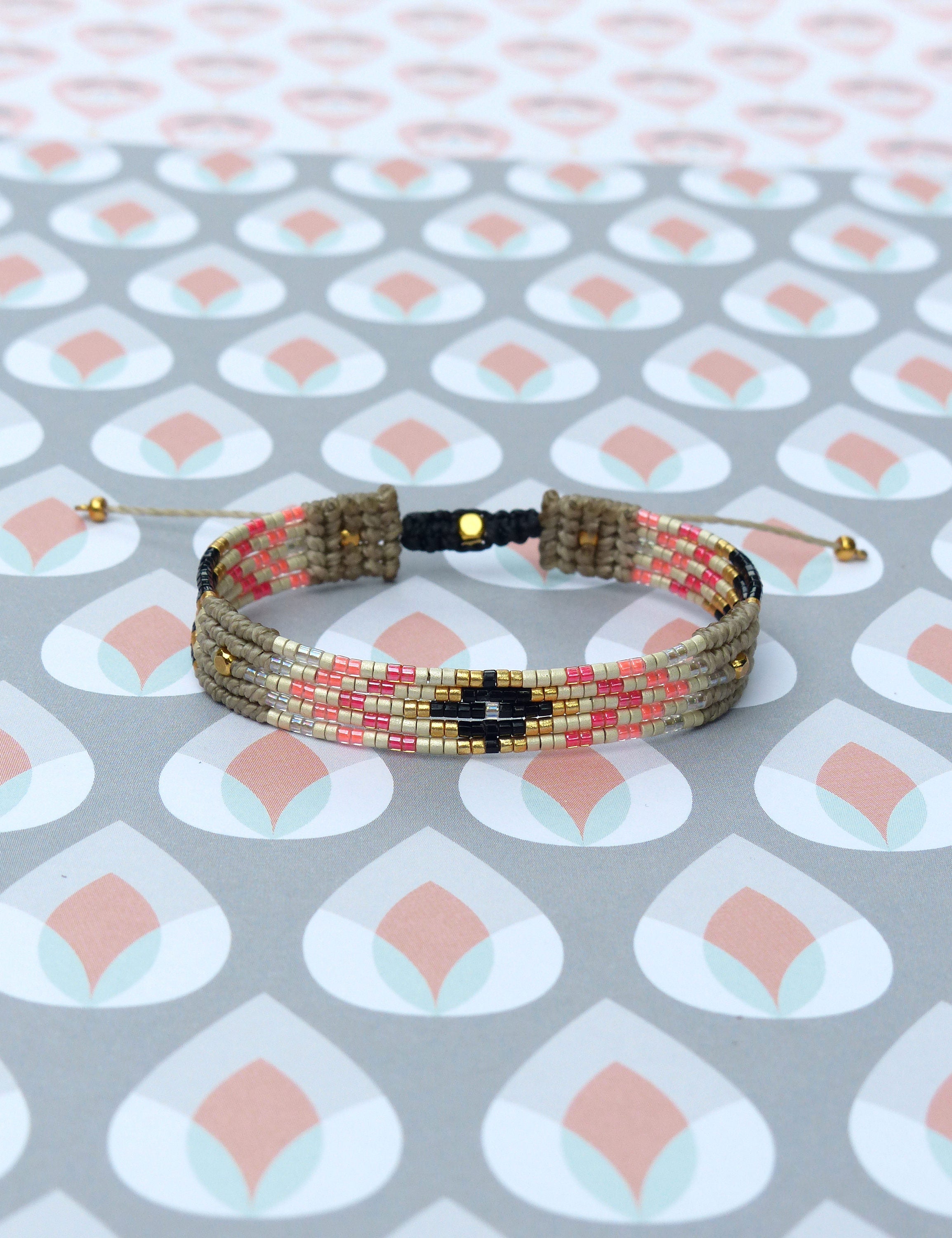 Peut inclure: Un bracelet tissé avec un motif géométrique. Le bracelet présente une base beige avec des rangées de perles roses, noires et dorées. Il est réglable avec des cordons beiges et des détails dorés.