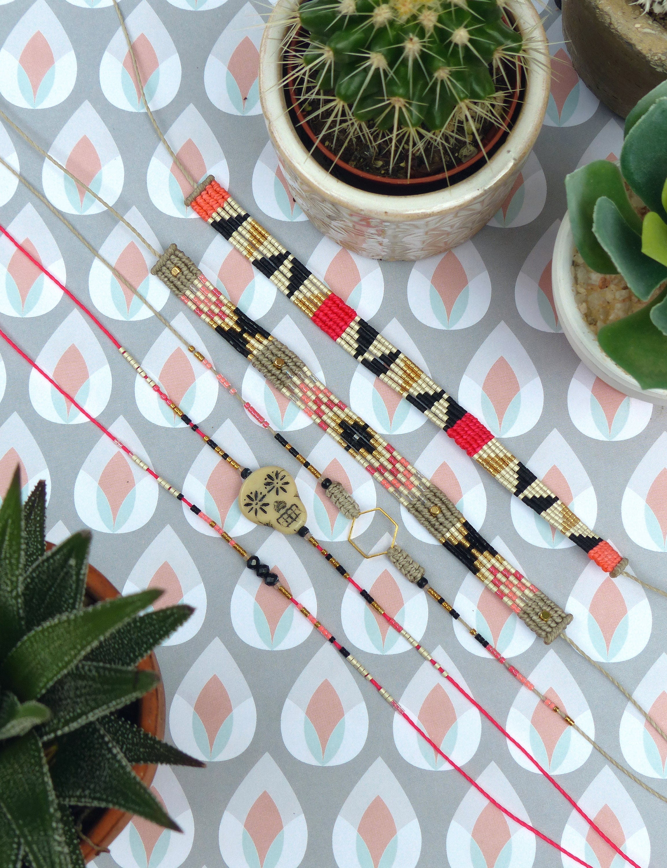 Peut inclure: Vue aérienne de colliers et bracelets faits à la main. Les bijoux présentent des motifs tissés et perlés en noir, rouge, or et rose. Un petit pendentif en forme de crâne est visible. L'arrière-plan comprend des plantes succulentes en pot et une surface à motifs.