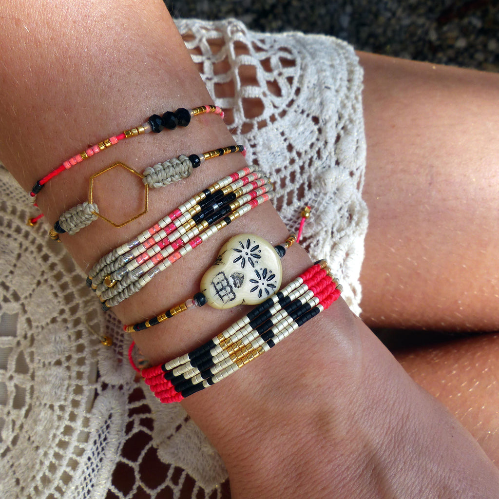 Peut inclure: Plusieurs bracelets de perles aux motifs et couleurs variés. Les bracelets comprennent des perles rouges, noires, dorées et blanches, avec un charm hexagonal et une perle décorative en forme de crâne. Les bracelets sont portés au poignet.