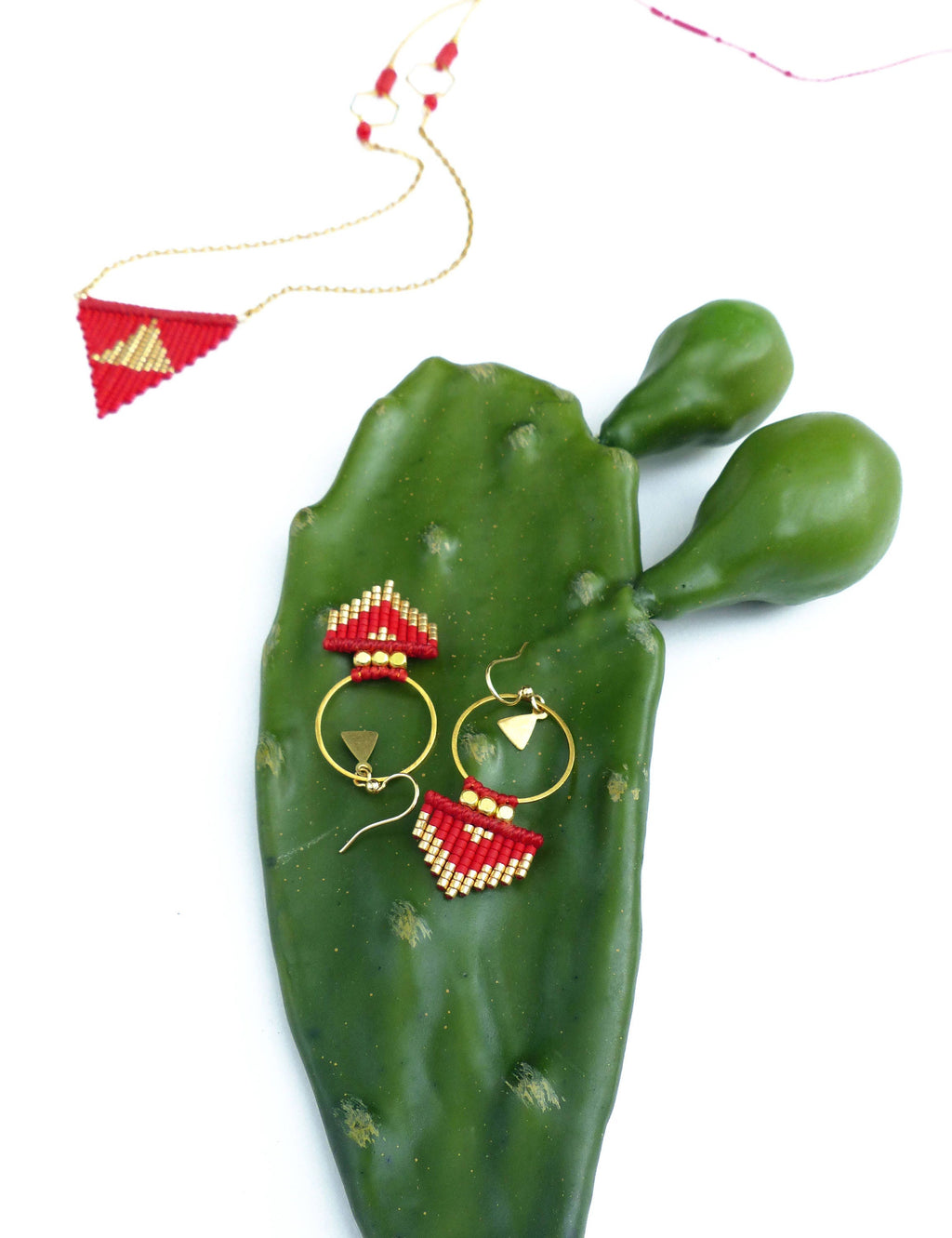 Peut inclure: Une paire de boucles d'oreilles créoles rouges et dorées avec un pendentif triangulaire suspendu à chaque créole. Les boucles d'oreilles sont présentées sur une feuille de cactus verte.