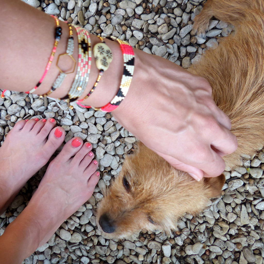 Peut inclure: Plusieurs bracelets colorés avec divers motifs, dont un charm en forme de crâne et des formes géométriques. L'image montre également les pieds d'une personne avec du vernis à ongles corail et un chien.