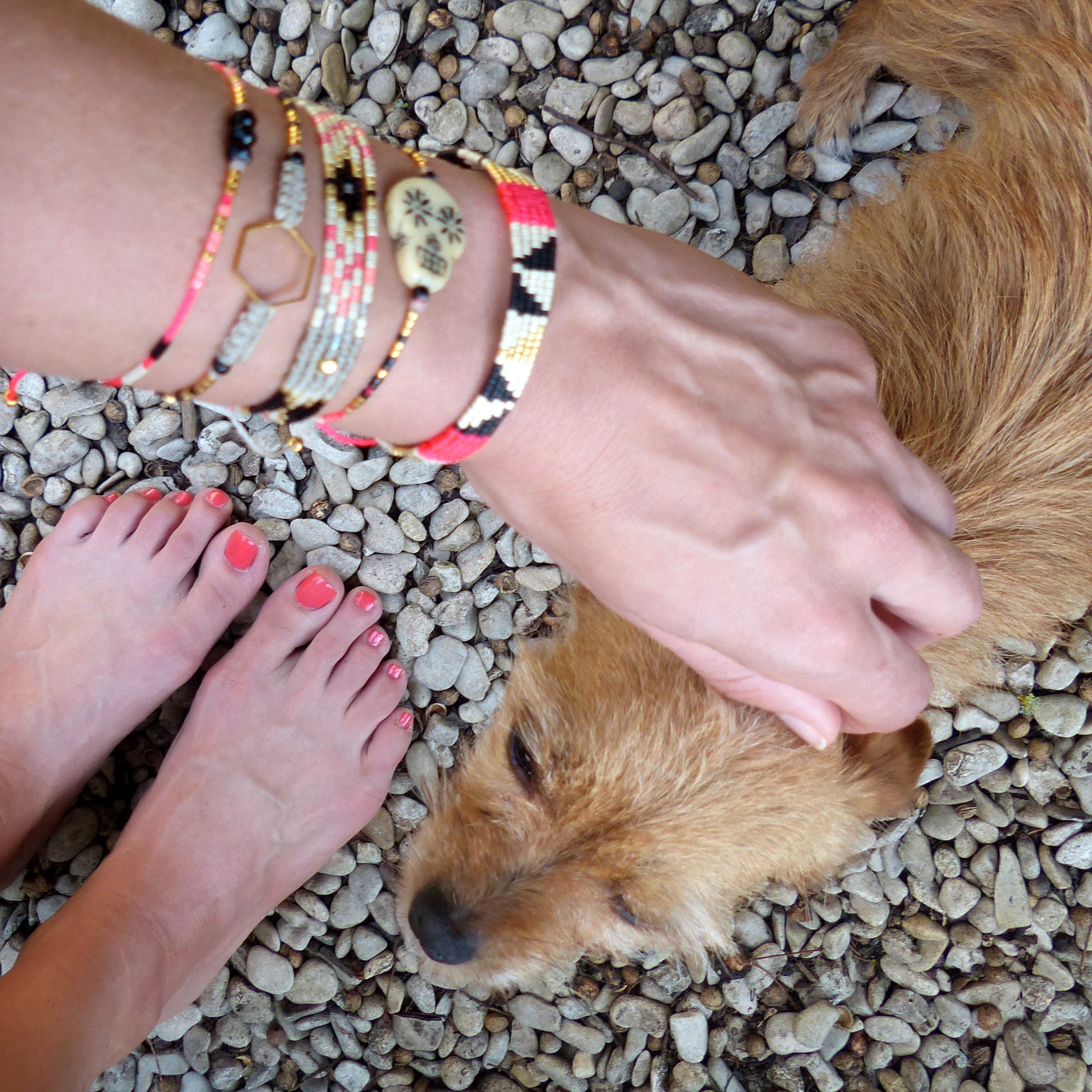 Peut inclure: Plusieurs bracelets colorés avec divers motifs, dont un charm en forme de crâne et des formes géométriques. L'image montre également les pieds d'une personne avec du vernis à ongles corail et un chien.