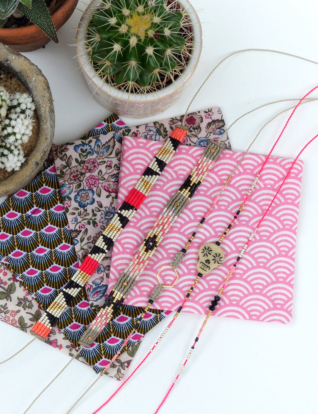 Peut inclure: Plusieurs bracelets de perles aux motifs complexes en noir, blanc, or et rouge sont présentés. Ils reposent sur des carrés de tissu à motifs, notamment des motifs floraux et vagues. Des cactus en pots sont visibles en arrière-plan.
