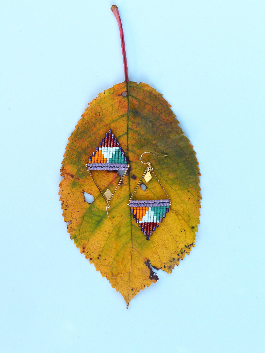 Peut inclure: Une paire de boucles d'oreilles triangulaires géométriques avec un cadre en fil d'or et des motifs de perles colorées. Les boucles d'oreilles sont présentées sur une feuille jaune.