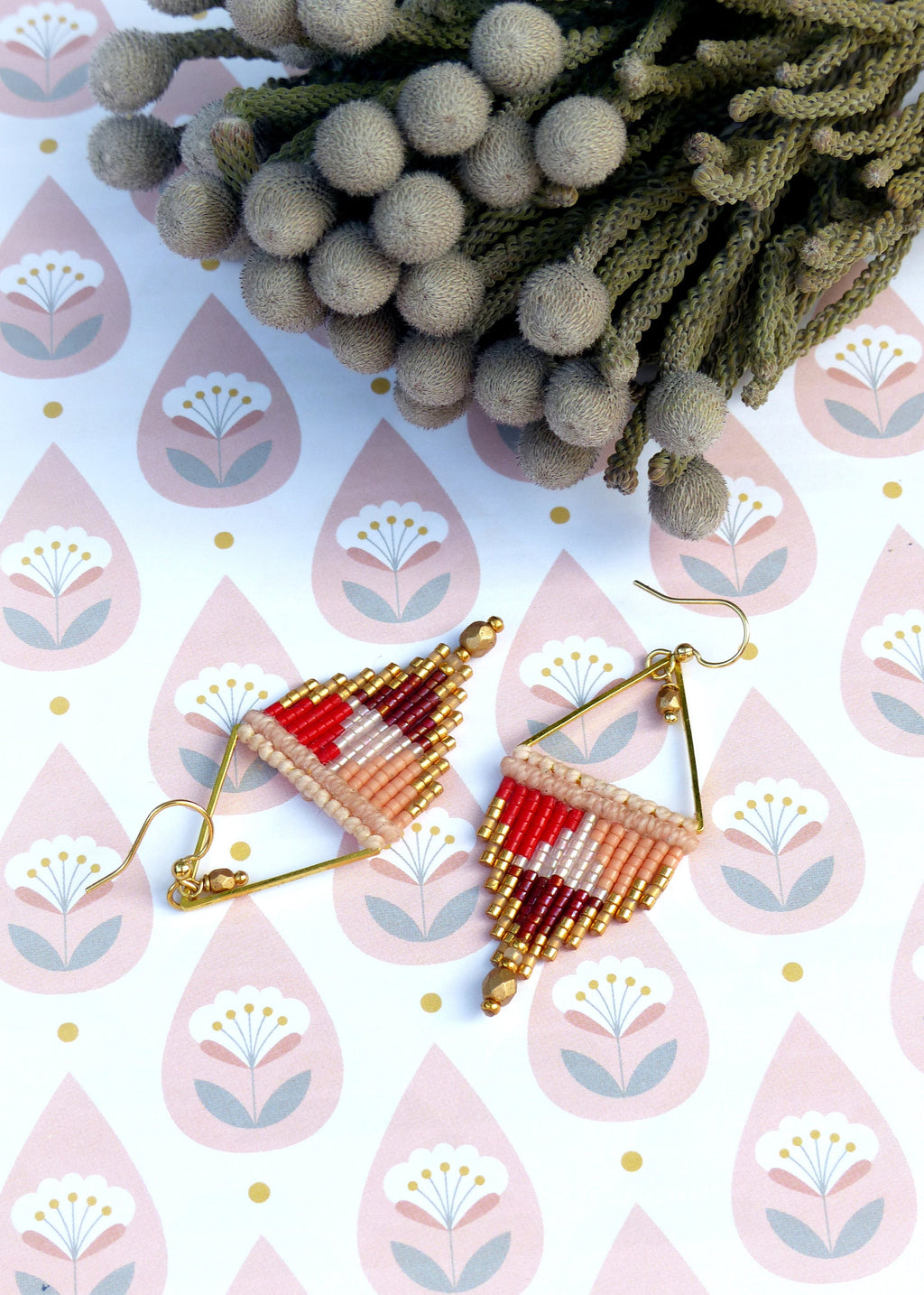 Peut inclure: Une paire de boucles d'oreilles triangulaires dorées avec des franges de perles rouges, roses et blanches. Les boucles d'oreilles sont sur un fond blanc avec un motif floral rose et gris.