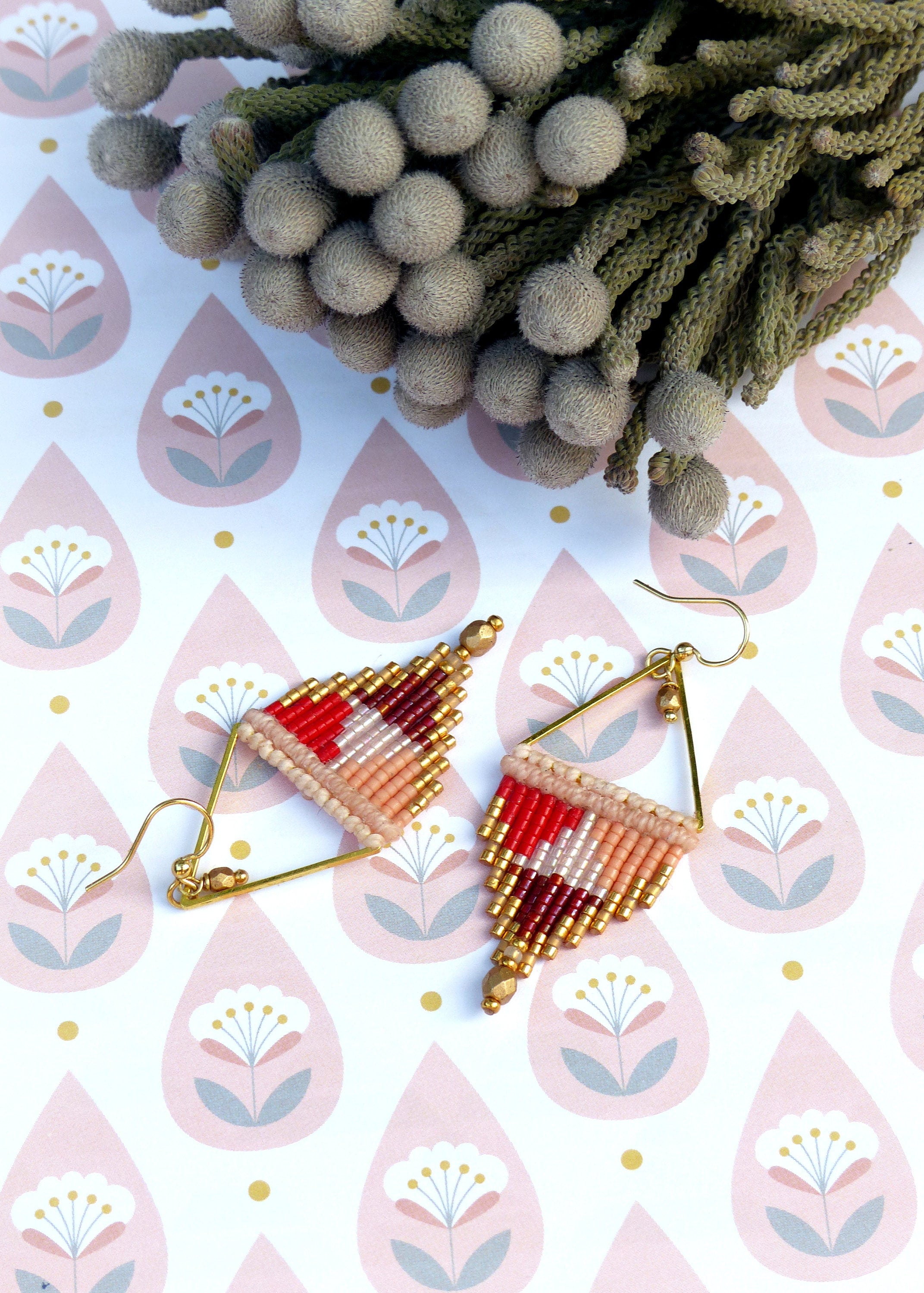 Peut inclure: Une paire de boucles d'oreilles triangulaires dorées avec des franges de perles rouges, roses et blanches. Les boucles d'oreilles sont sur un fond blanc avec un motif floral rose et gris.