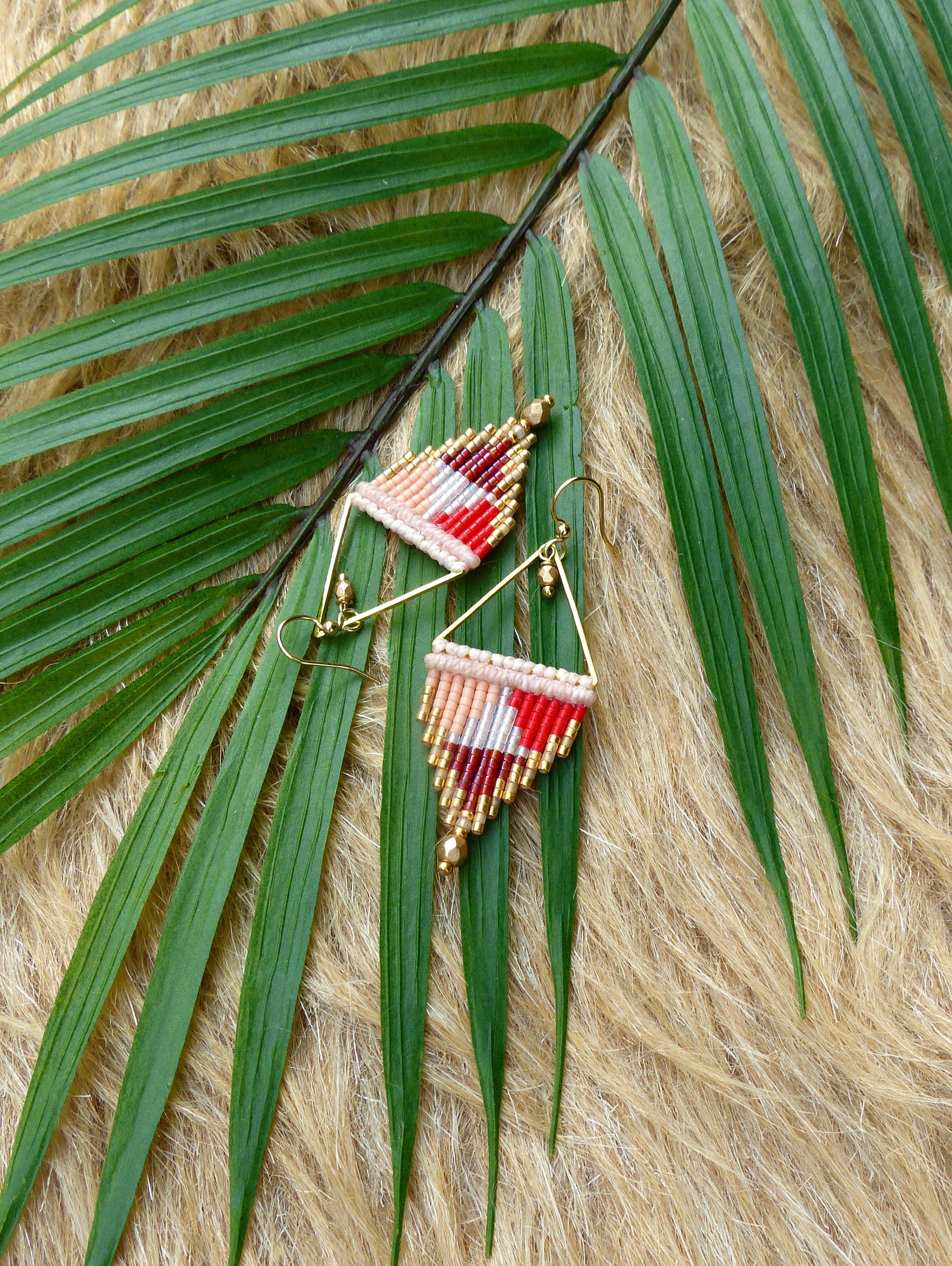 Peut inclure: Une paire de boucles d'oreilles en forme de triangle avec un motif géométrique de perles rouges, blanches et roses. Les boucles d'oreilles sont faites de fil de couleur or et ont une perle pendante en bas.