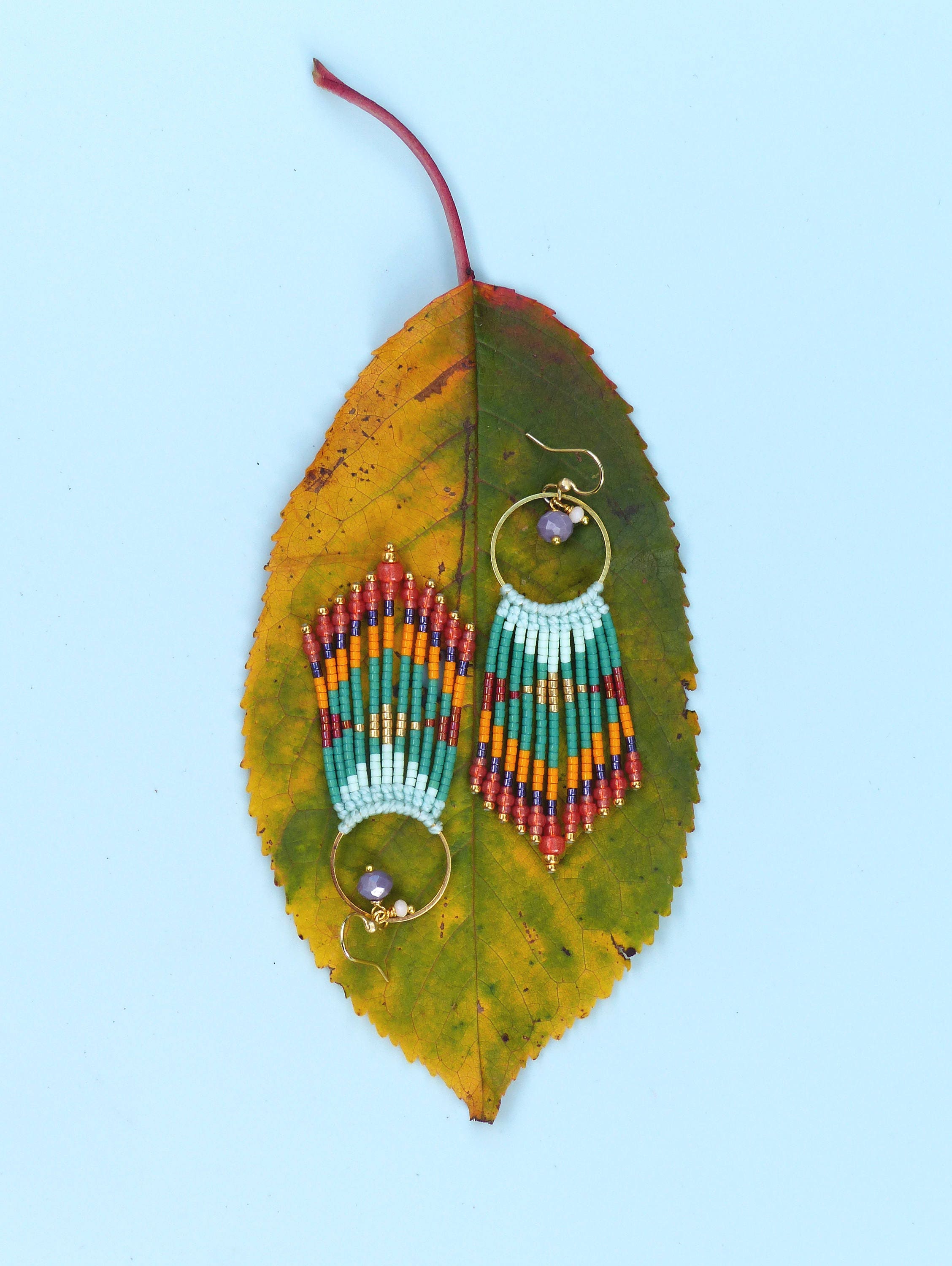 Peut inclure: Une paire de longues boucles d'oreilles en perles colorées avec des cercles dorés et des perles violettes qui pendent du bas. Les boucles d'oreilles sont présentées sur une feuille jaune et verte.