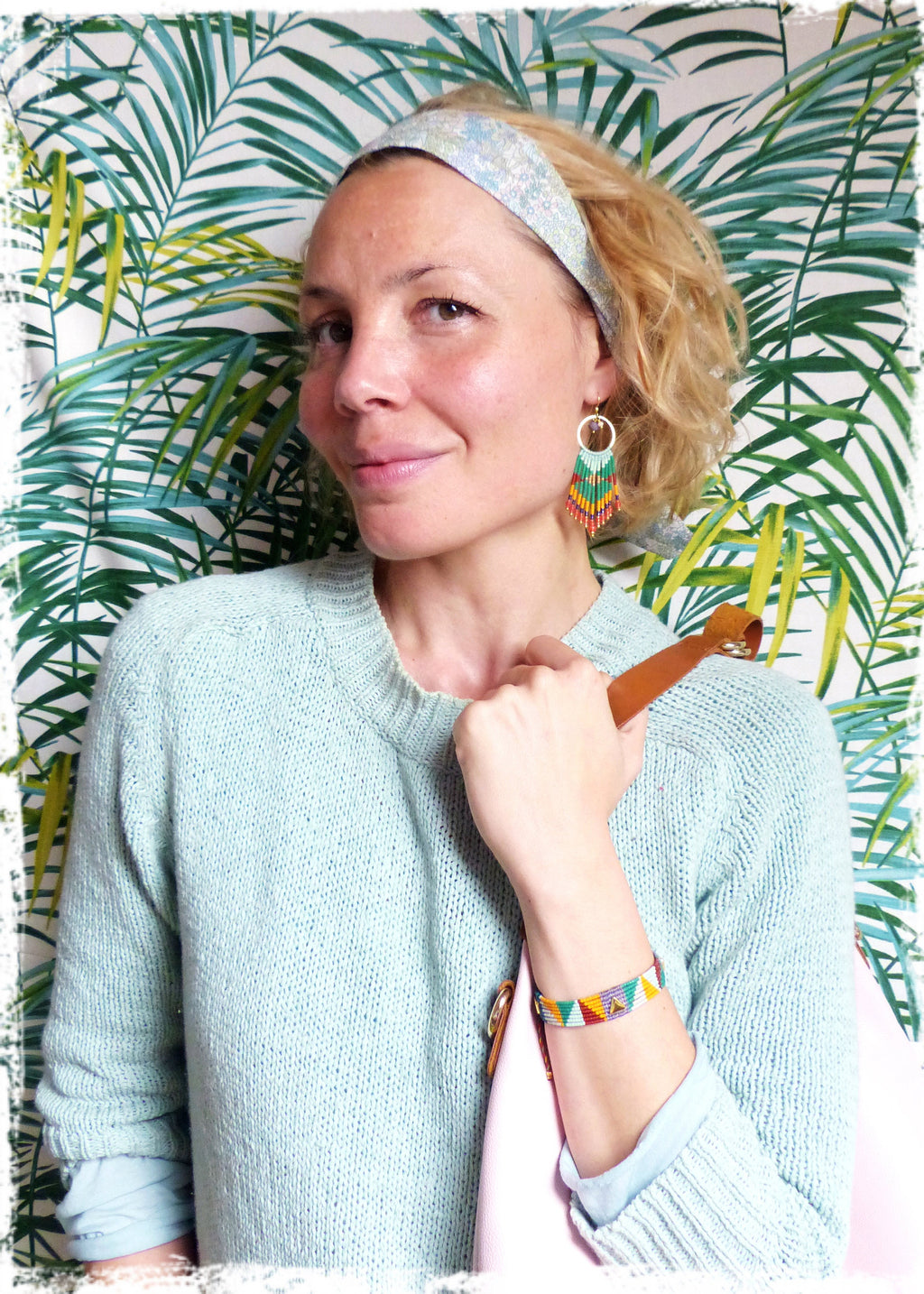 Peut inclure: Une femme portant un pull bleu clair avec une bandoulière en cuir marron sur l'épaule. Elle porte un bandeau bleu clair à motifs blancs et bleus, et une paire de boucles d'oreilles en perles colorées. Elle porte également un bracelet en perles colorées au poignet.
