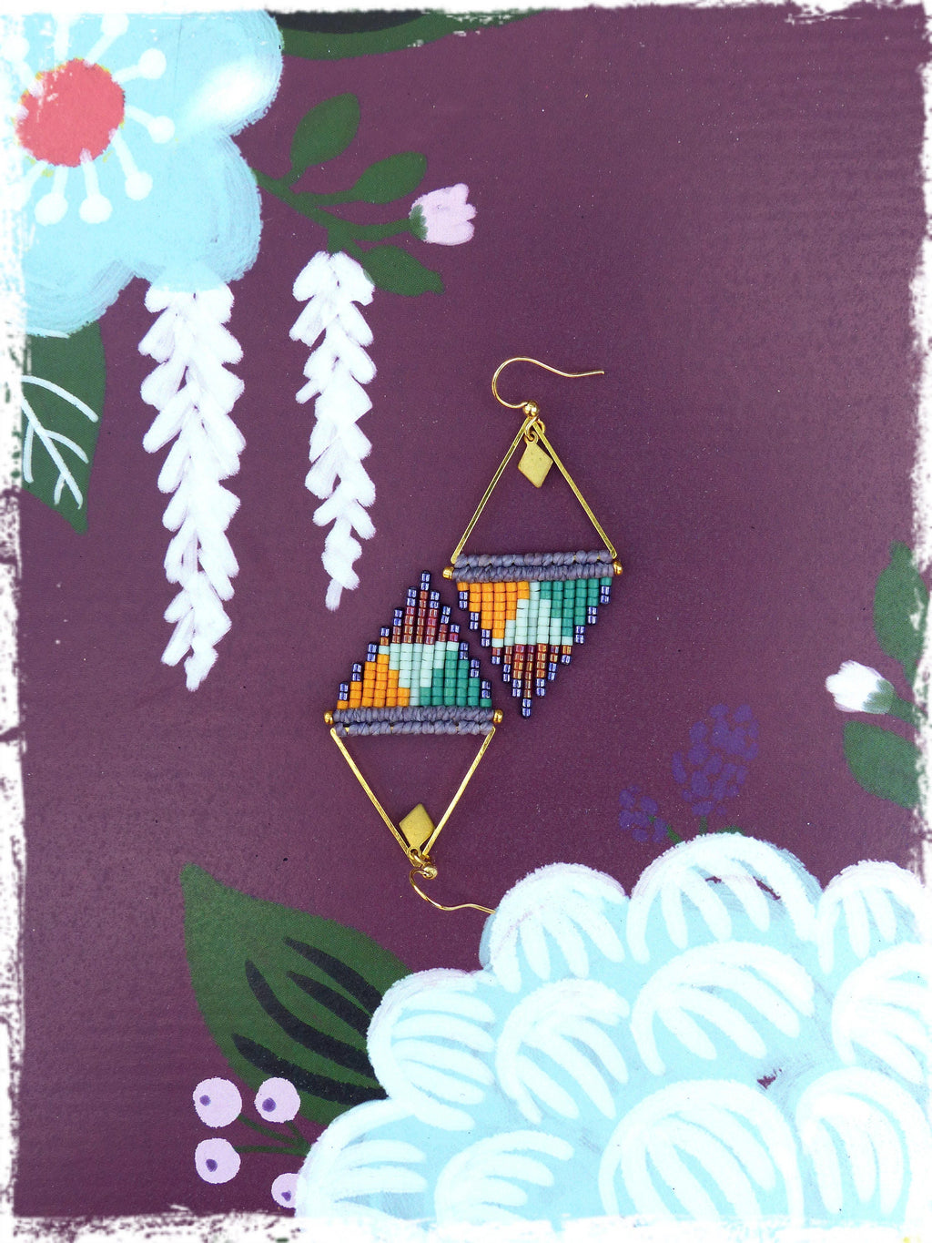 Peut inclure: Une paire de boucles d'oreilles géométriques dorées de forme triangulaire. Les boucles d'oreilles sont faites de perles colorées dans des tons d'orange, de vert, de bleu et de violet. Les boucles d'oreilles sont suspendues sur un fond violet avec des fleurs peintes.