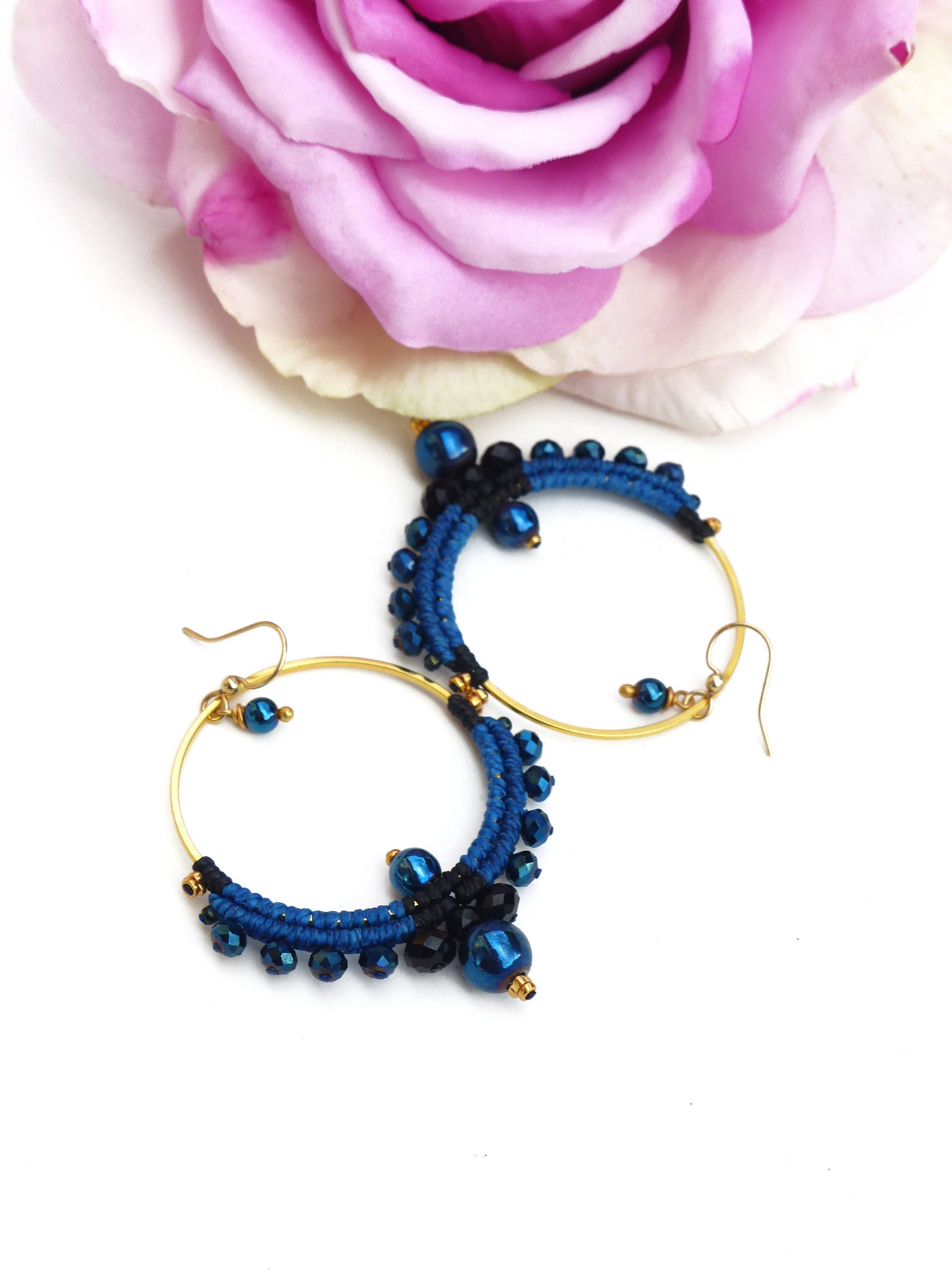 Peut inclure: Une paire de boucles d'oreilles créoles dorées avec des perles bleues et noires. Les boucles d'oreilles sont réalisées à l'aide d'une technique de macramé et présentent un groupe de perles en bas.